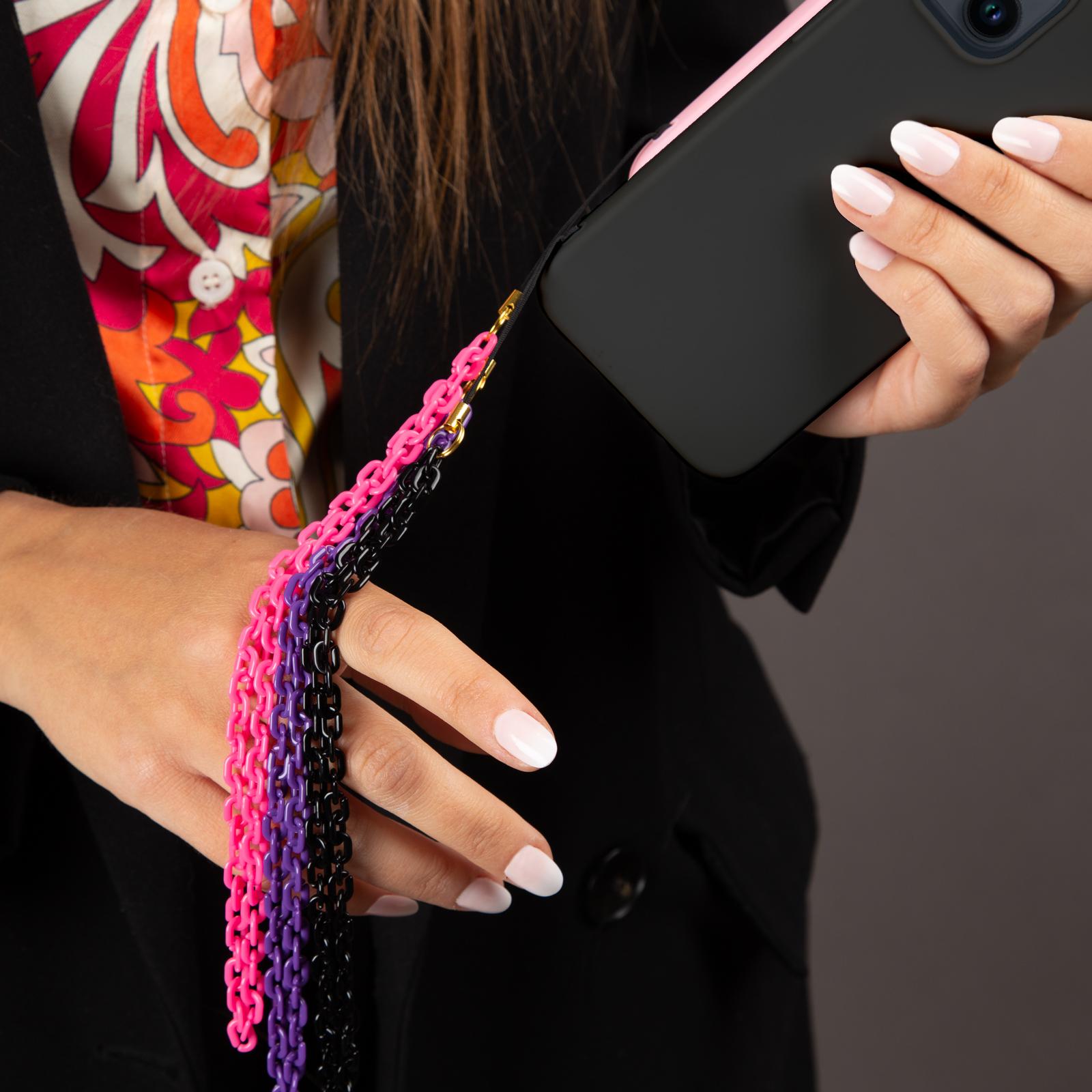 Una persona sostiene un teléfono negro con funda rosa y una cadena de eslabones rosa, morados y negros.