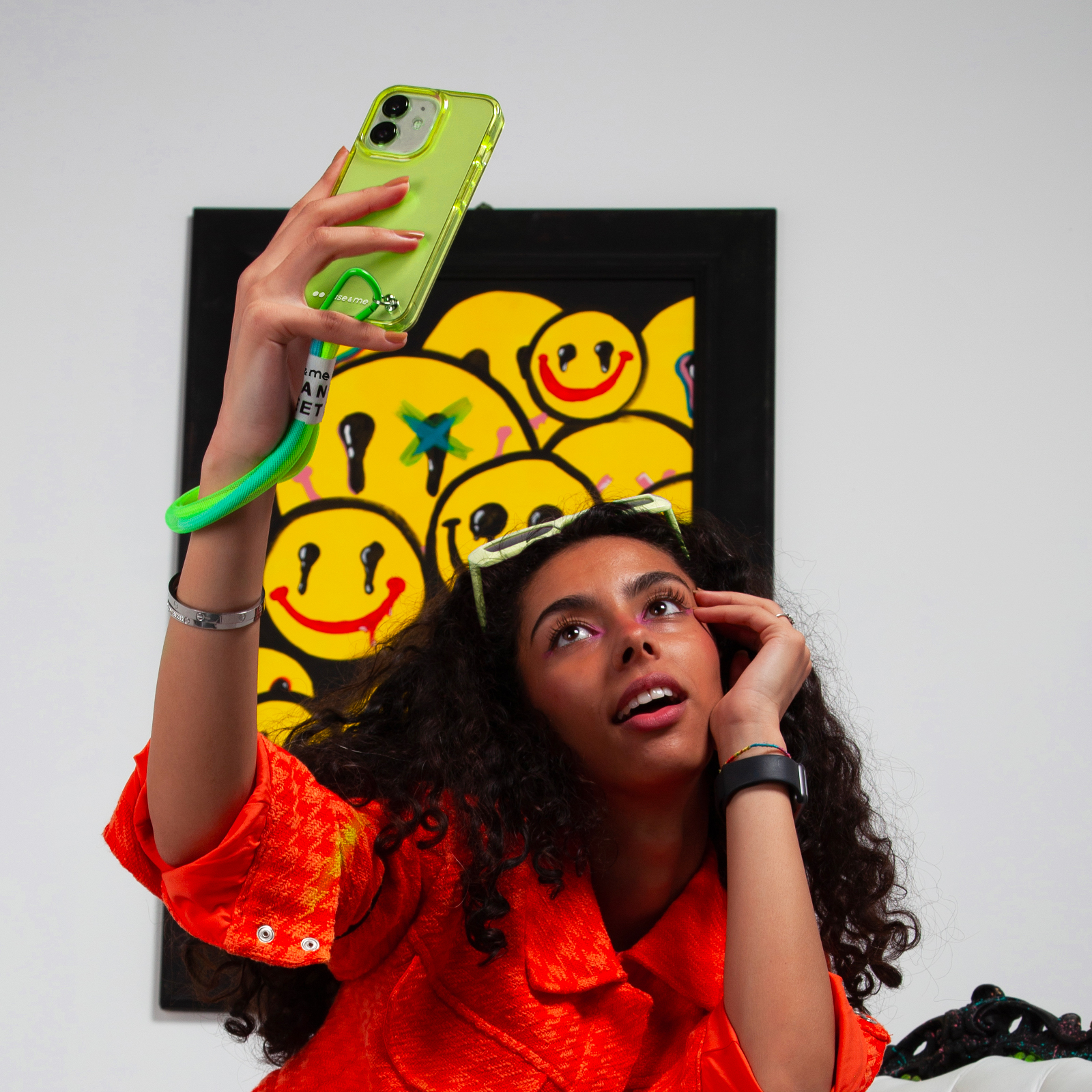 Una mujer se toma una selfie con un teléfono verde neón, con una pintura de caras sonrientes detrás.