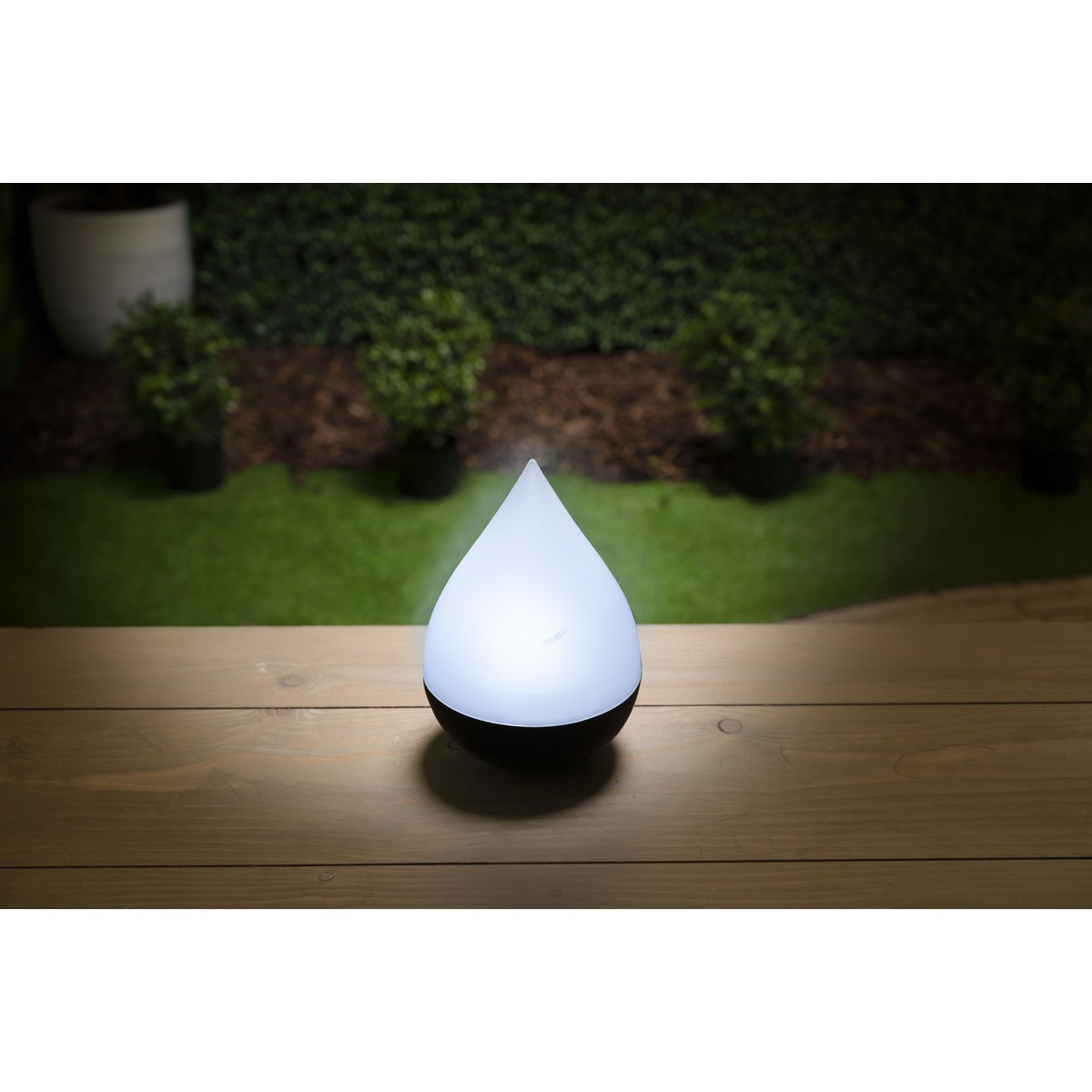 A white teardrop-shaped lantern glows on a wooden deck, with a blurred garden background.