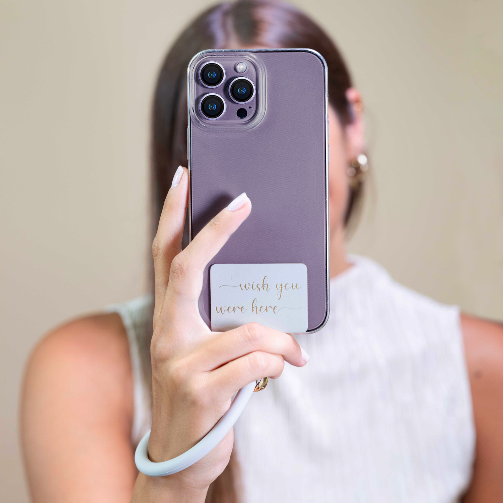 Una mano sostiene un smartphone morado con cámaras y una pegatina que dice 'wish you were here'.