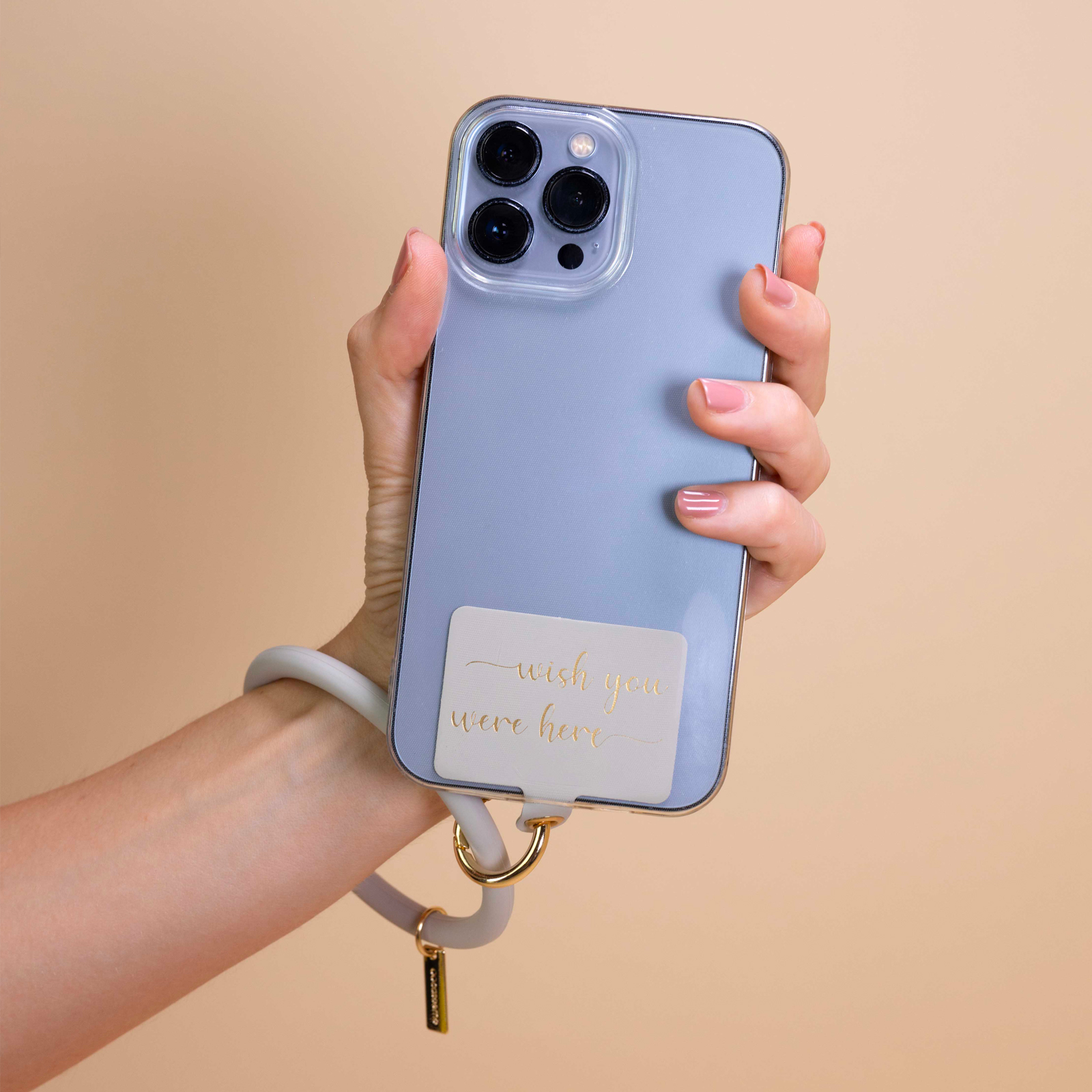 Una mano sostiene un teléfono azul con correa de muñeca. Texto dorado dice "wish you were here".