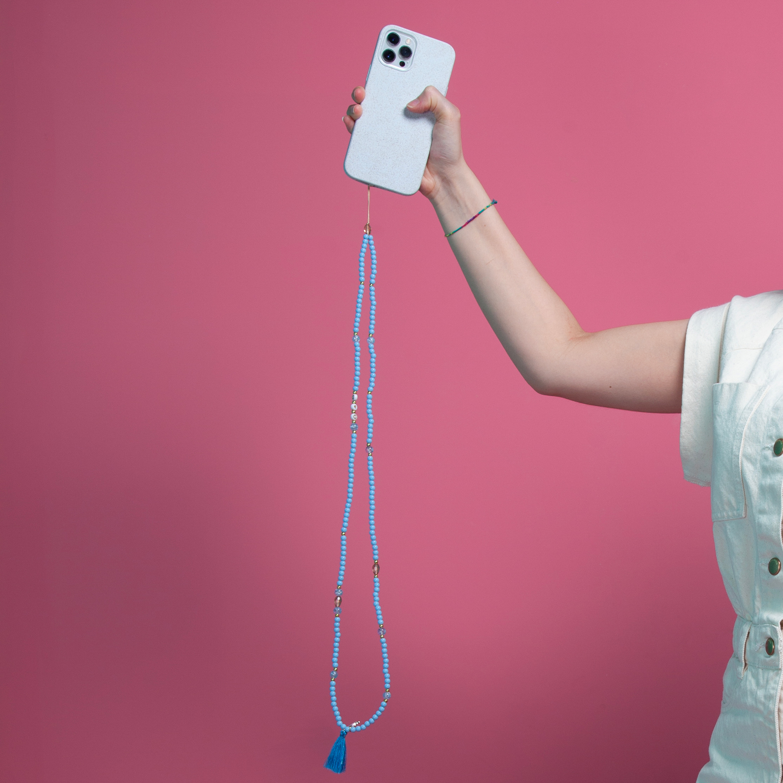Una mano sostiene un teléfono azul claro con una cadena de cuentas azules y un charm de borla.