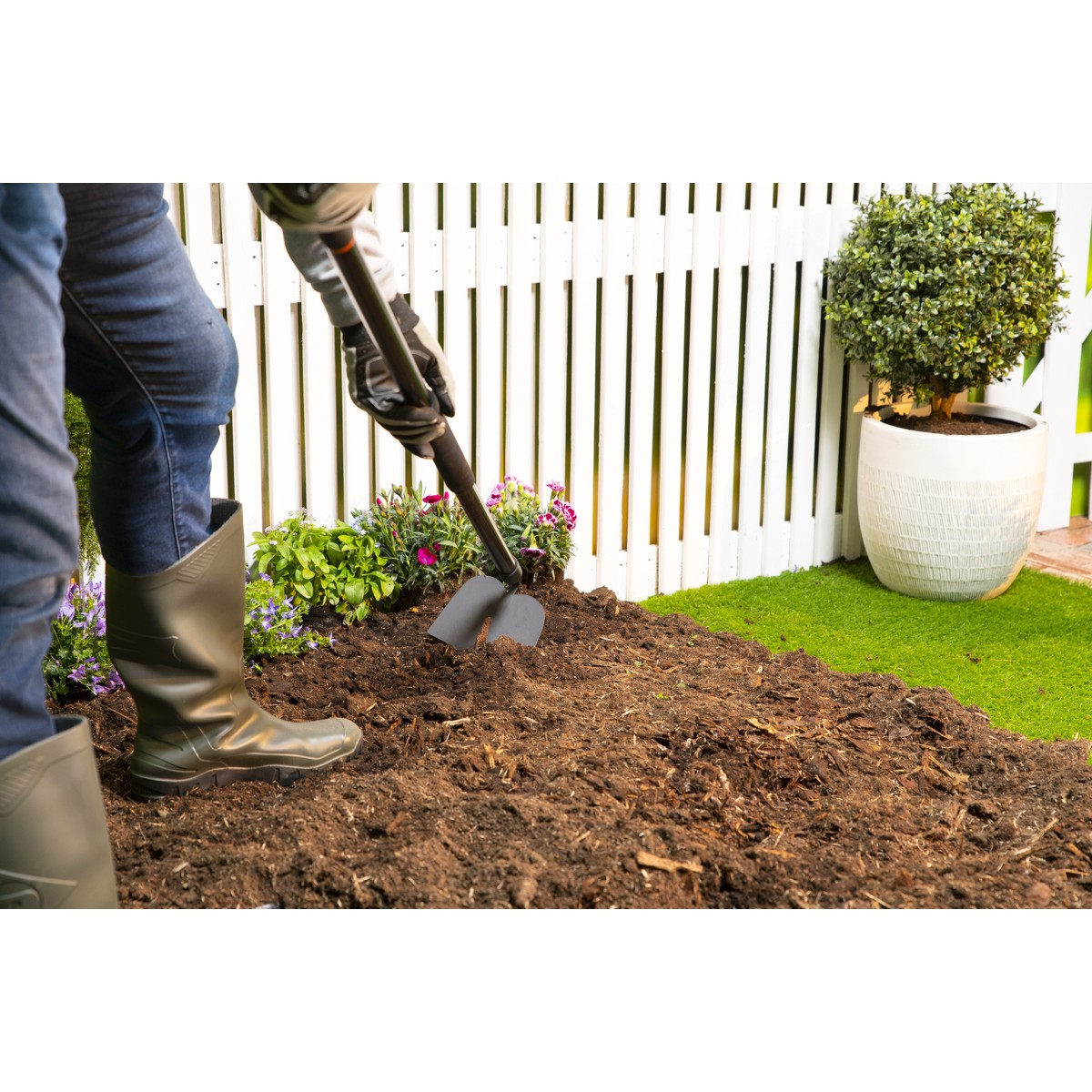 Person digging in soil with a spade near a white fence and potted plant.