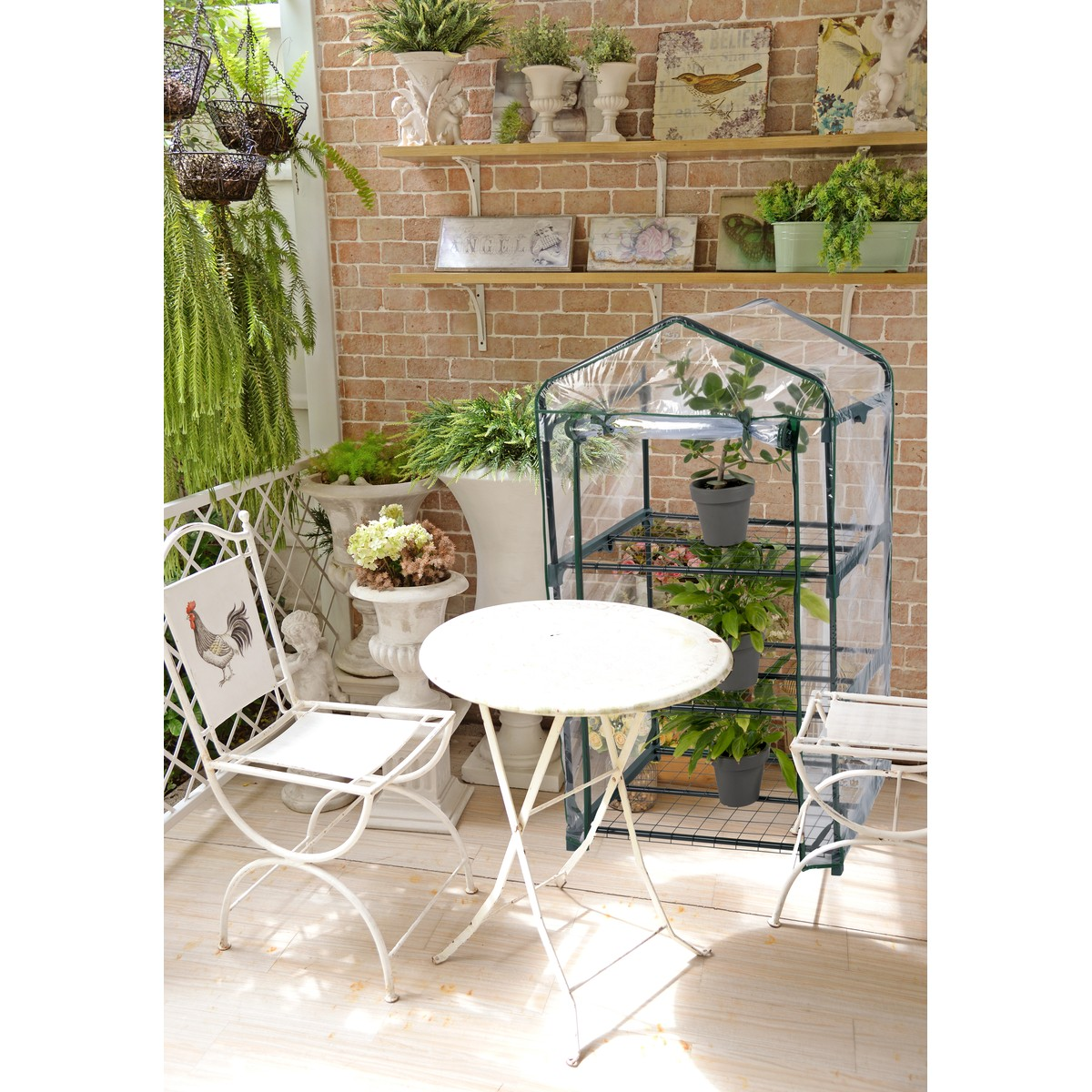 A white garden table and chair with a rooster motif in front of a greenhouse with plants.