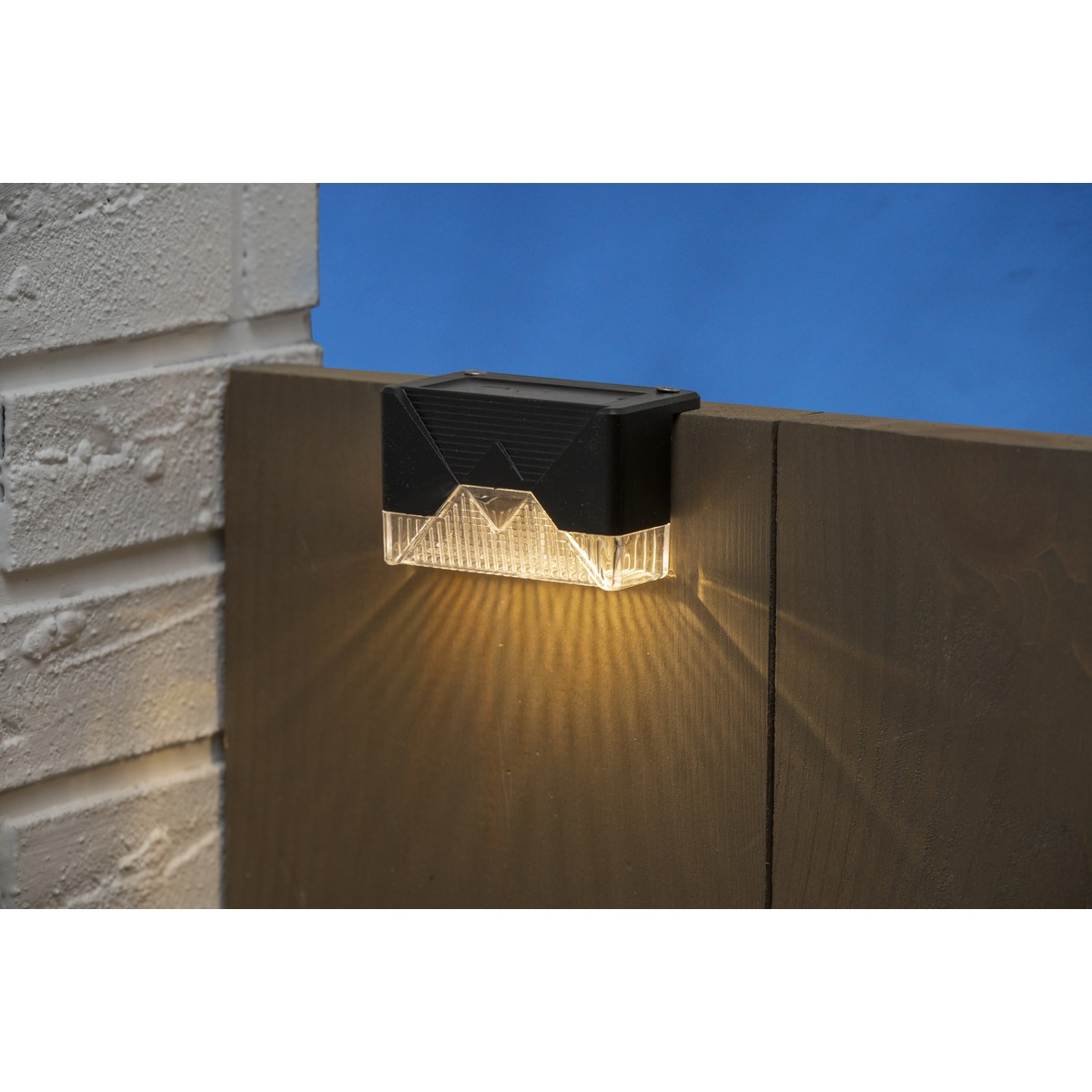 A black solar fence light shines yellow on a wooden post against a blue sky.
