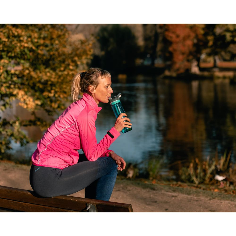 Frau in pinker Jacke trinkt aus blauer Wasserflasche auf einer Parkbank am See.