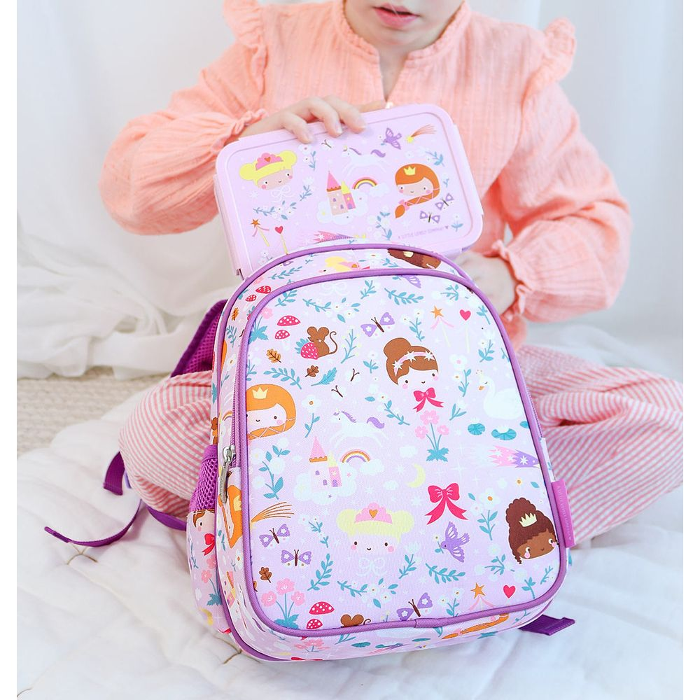Ein Kind in einem pfirsichfarbenen Oberteil hält eine Brotdose, die zu einem pinkfarbenen Rucksack mit Prinzessinnen- und Blumenmotiven passt.