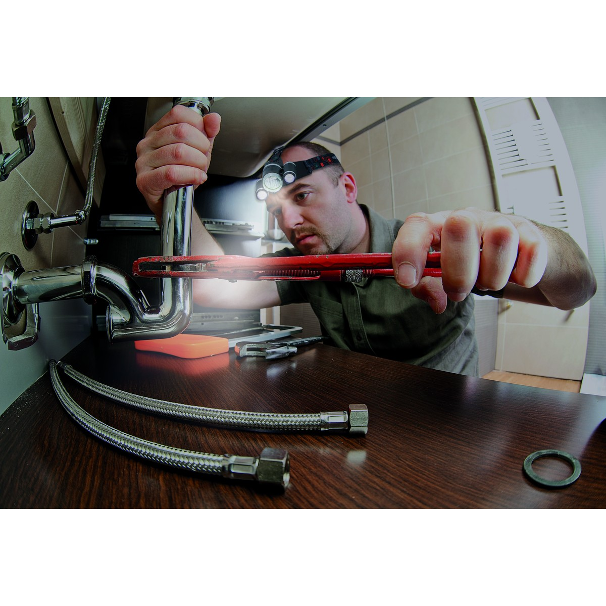 Plumber with headlamp repairs pipes using a wrench on a wooden surface.