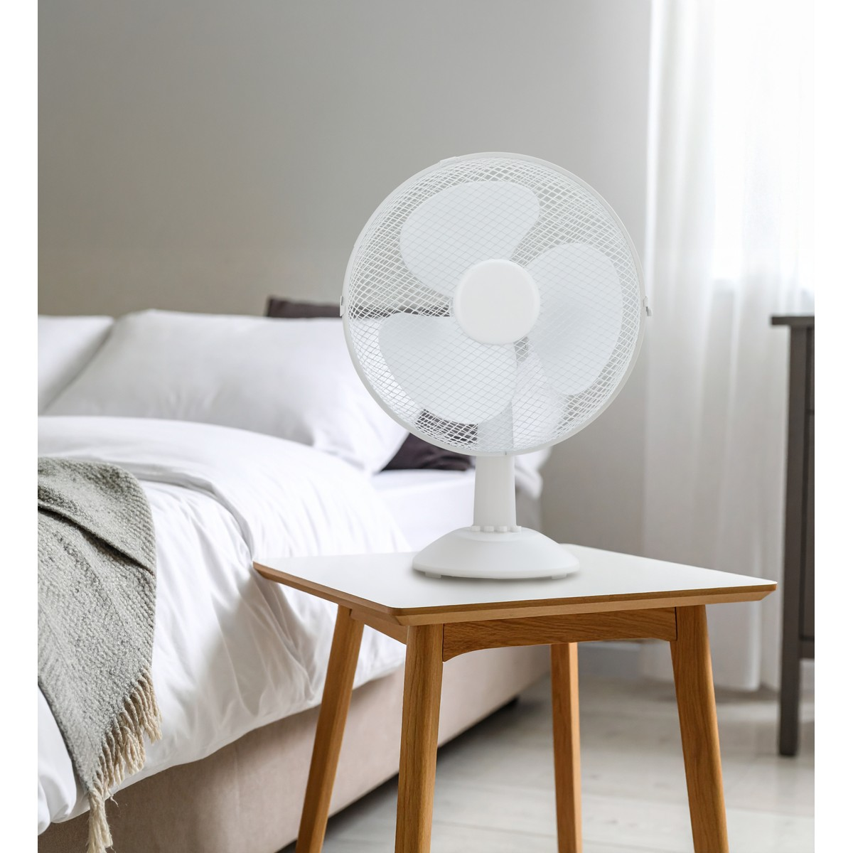Ein weißer Ventilator steht auf einem Beistelltisch neben einem Bett mit weißen Bettwäsche.