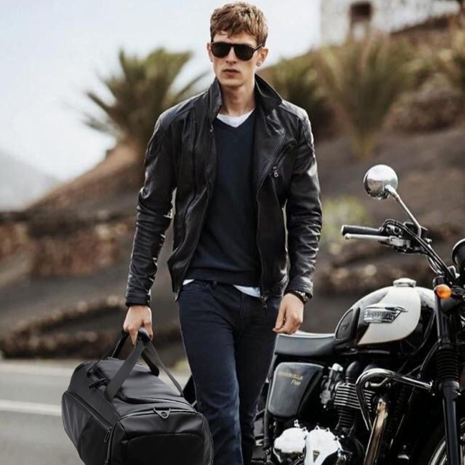 Mann mit Lederjacke und Tasche geht neben Motorrad in sonniger Landschaft.