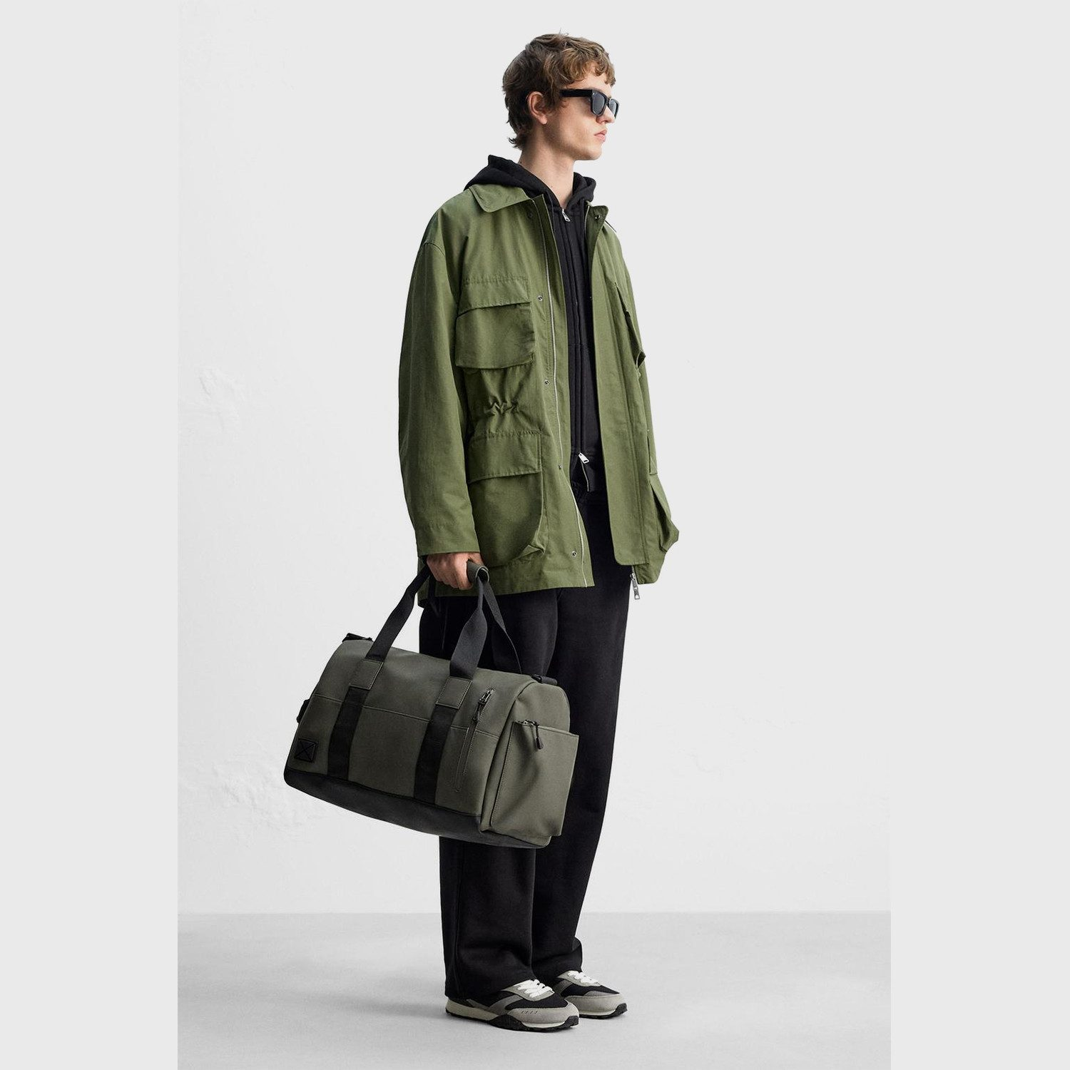 Ein Mann mit Sonnenbrille trägt eine grüne Jacke und hält eine dunkelgrüne Tasche.