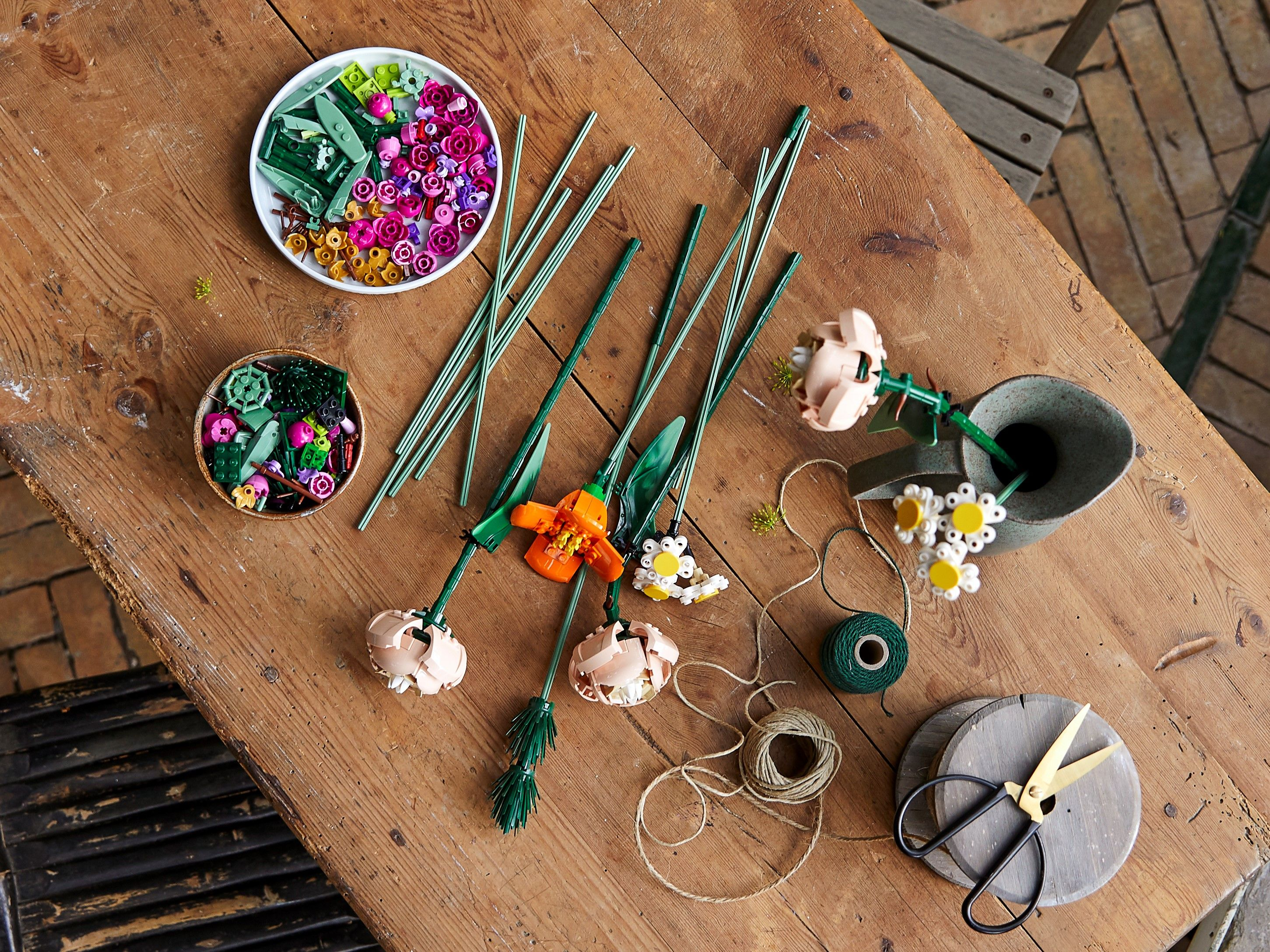 Colorful Lego flower building parts with stems, twine, and scissors on a wooden table.