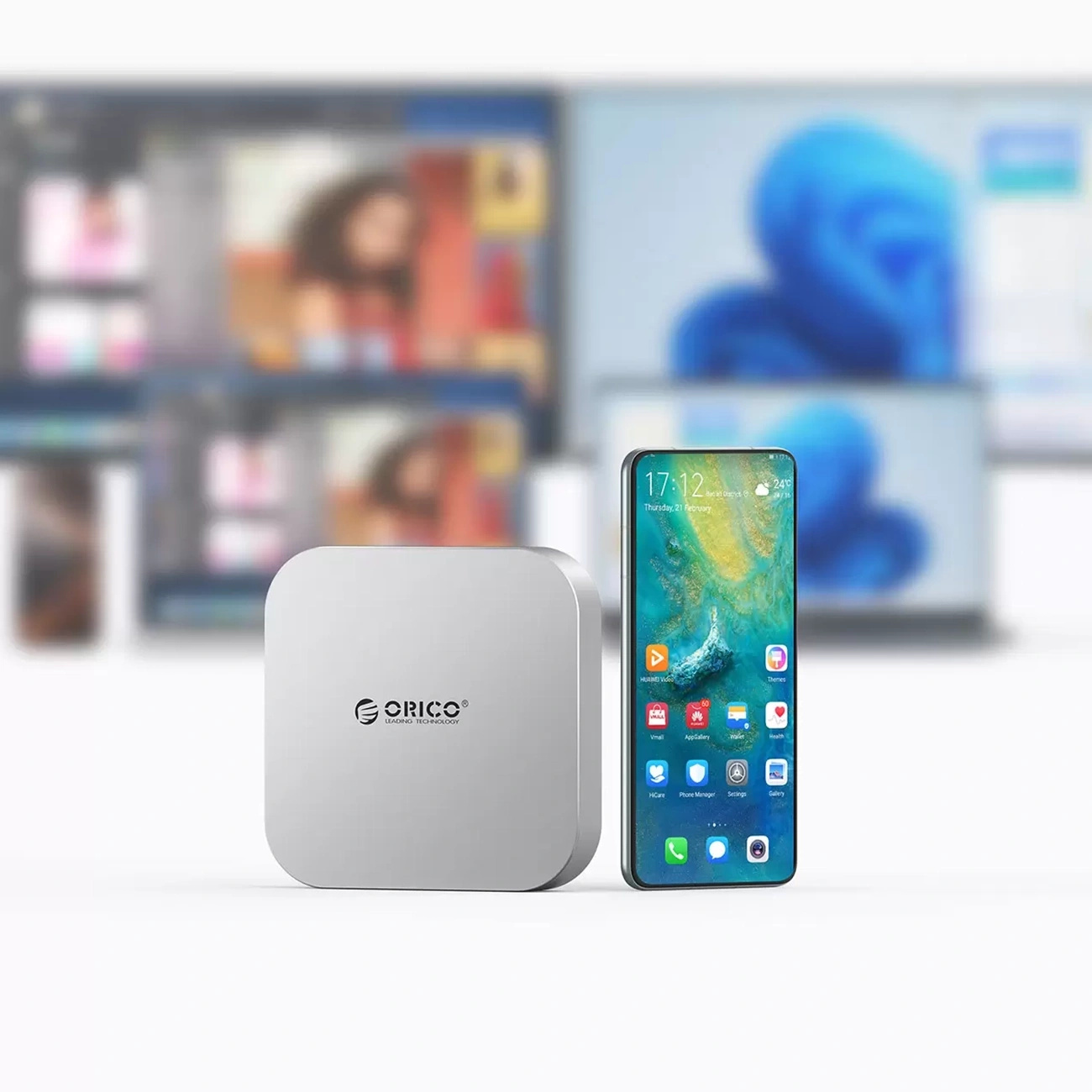 Srebrne urządzenie ORICO i smartfon na białej powierzchni z rozmytymi ekranami w tle.