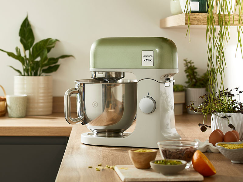 Una impastatrice Kenwood verde e bianca su un bancone di legno con ingredienti.