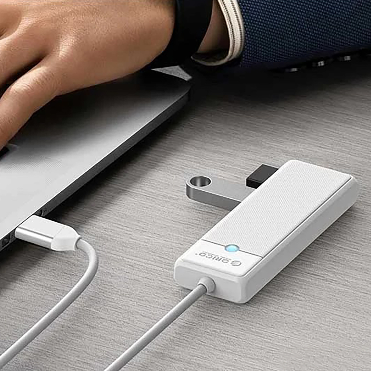 Dłoń osoby obok srebrnego laptopa, z podłączonym białym hubem USB-C, posiadającym niebieską lampkę kontrolną.