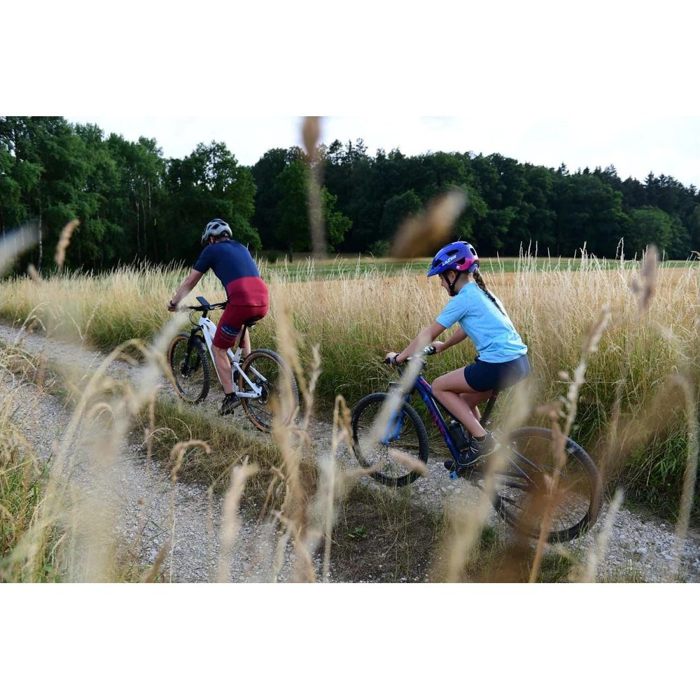 Zwei Radfahrer fahren auf einem Feldweg durch trockenes Gras, Bäume im Hintergrund.