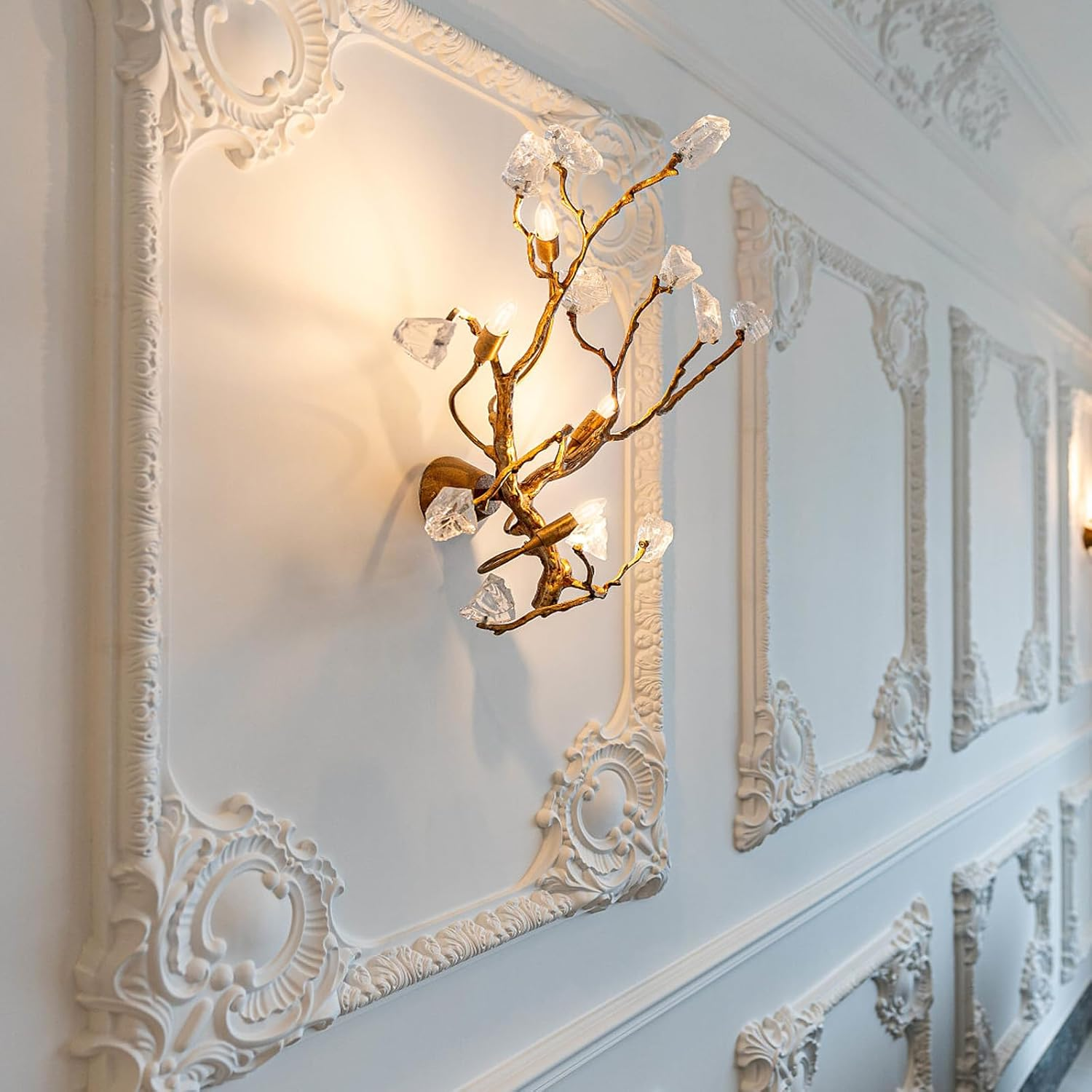 Ein goldener Wandleuchter in Astform mit Kristallblättern an einer weißen, verzierten Wand.