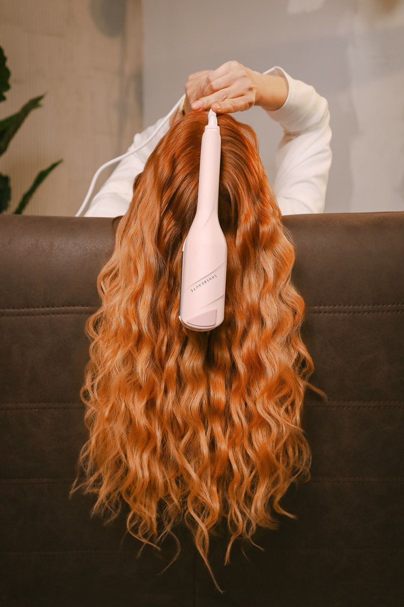Eine Person mit langen, welligen, orangefarbenen Haaren hält einen rosa Lockenstab.