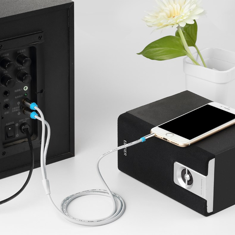 A white audio cable connects a smartphone to a black speaker. Another cable connects to a larger speaker.