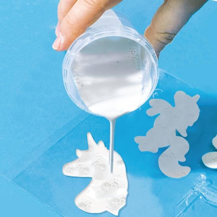 Una mano rovescia una tazza trasparente con liquido bianco su uno stampo a forma di unicorno su superficie blu.