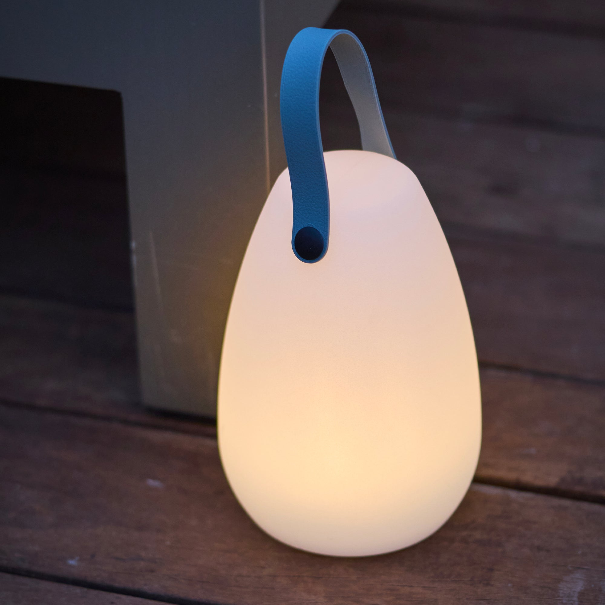 Eine weiße, eiförmige Lampe mit blauem Henkel steht auf Holzbrettern.