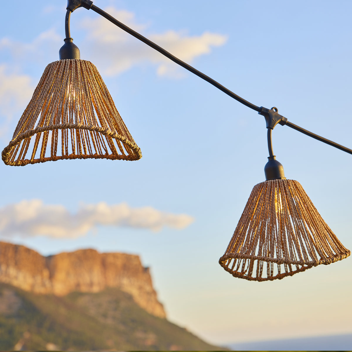 Zwei geflochtene Lampenschirme hängen an einem Kabel vor einem blauen Himmel mit Wolken und Berg im Hintergrund.