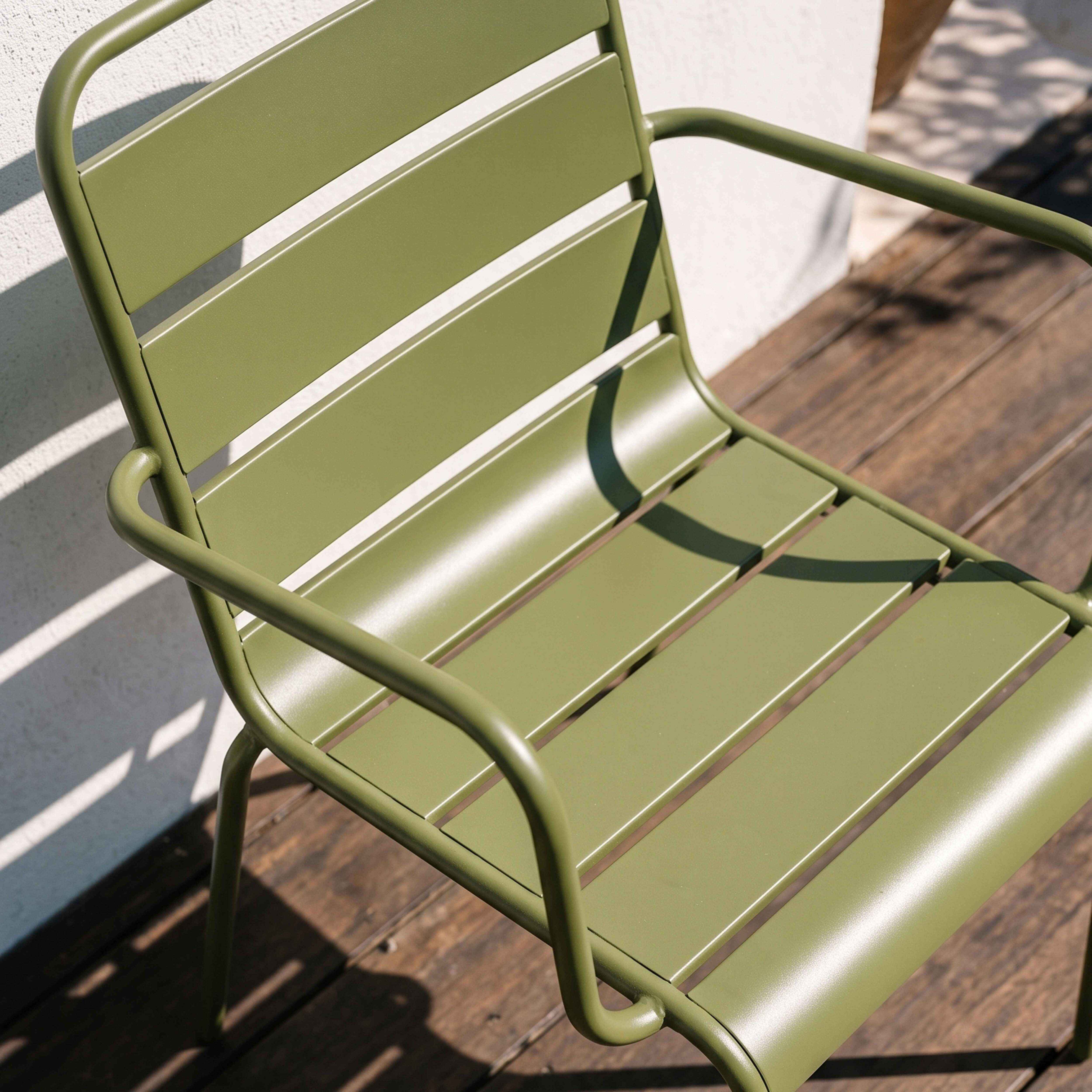 Ein grüner Metallstuhl steht auf einer Holzterrasse. Schatten fallen auf die Wand.