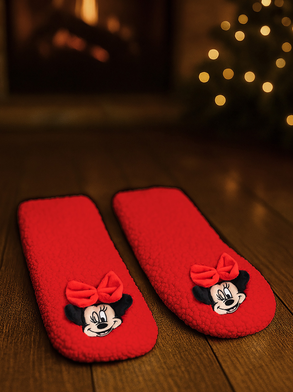 Rote Hausschuhe mit Minnie Maus-Motiv auf Holzboden, Kamin und Lichterkette im Hintergrund.