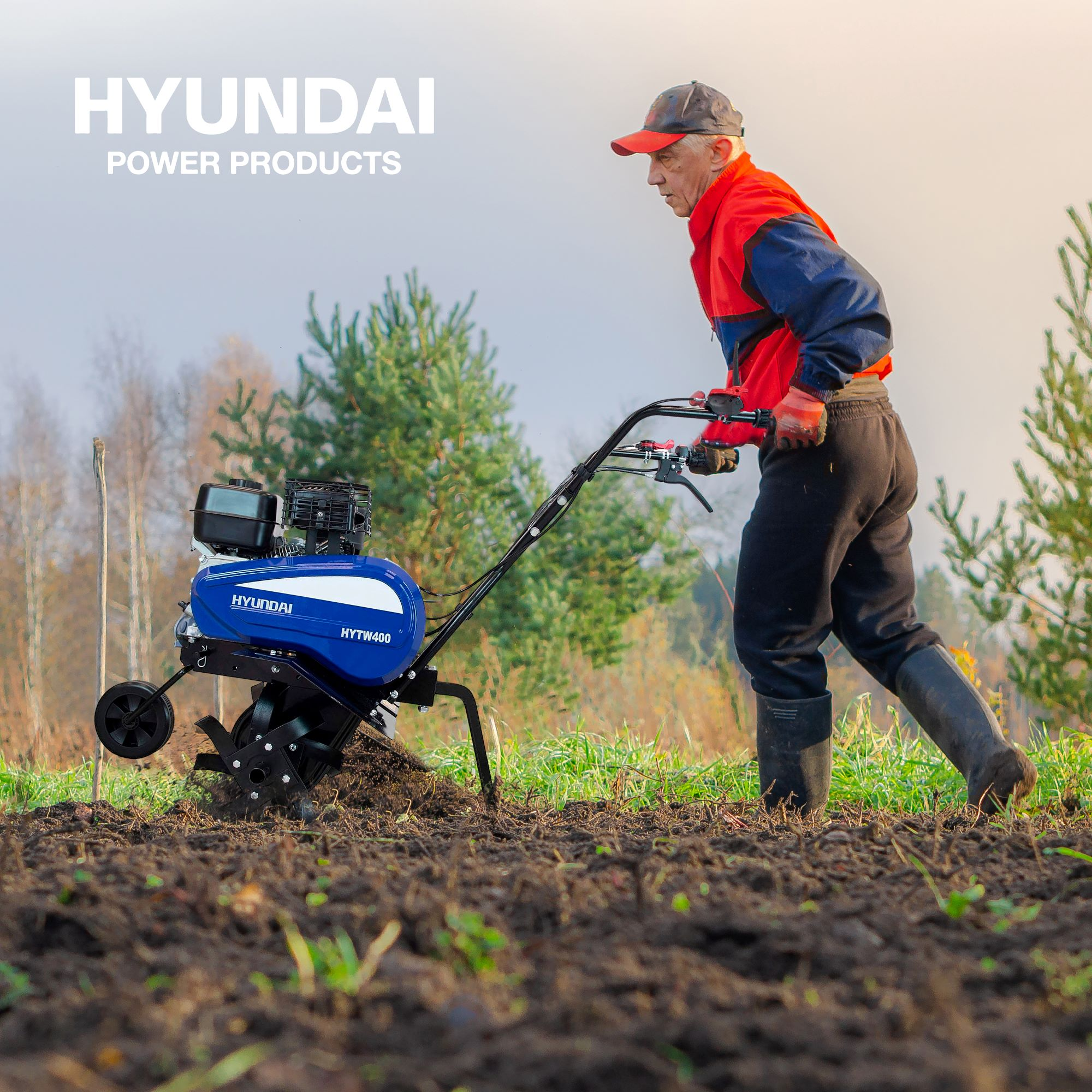Un hombre mayor opera una motoazada Hyundai azul, labrando la tierra en un campo con árboles.