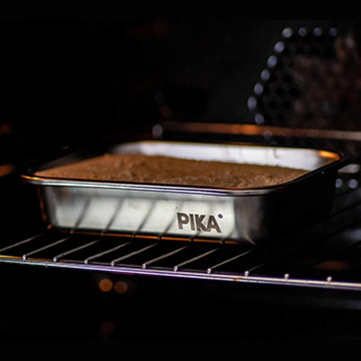 Prostokątne ciasto piecze się w srebrnej formie na ruszcie piekarnika. Forma ma logo PIKA.