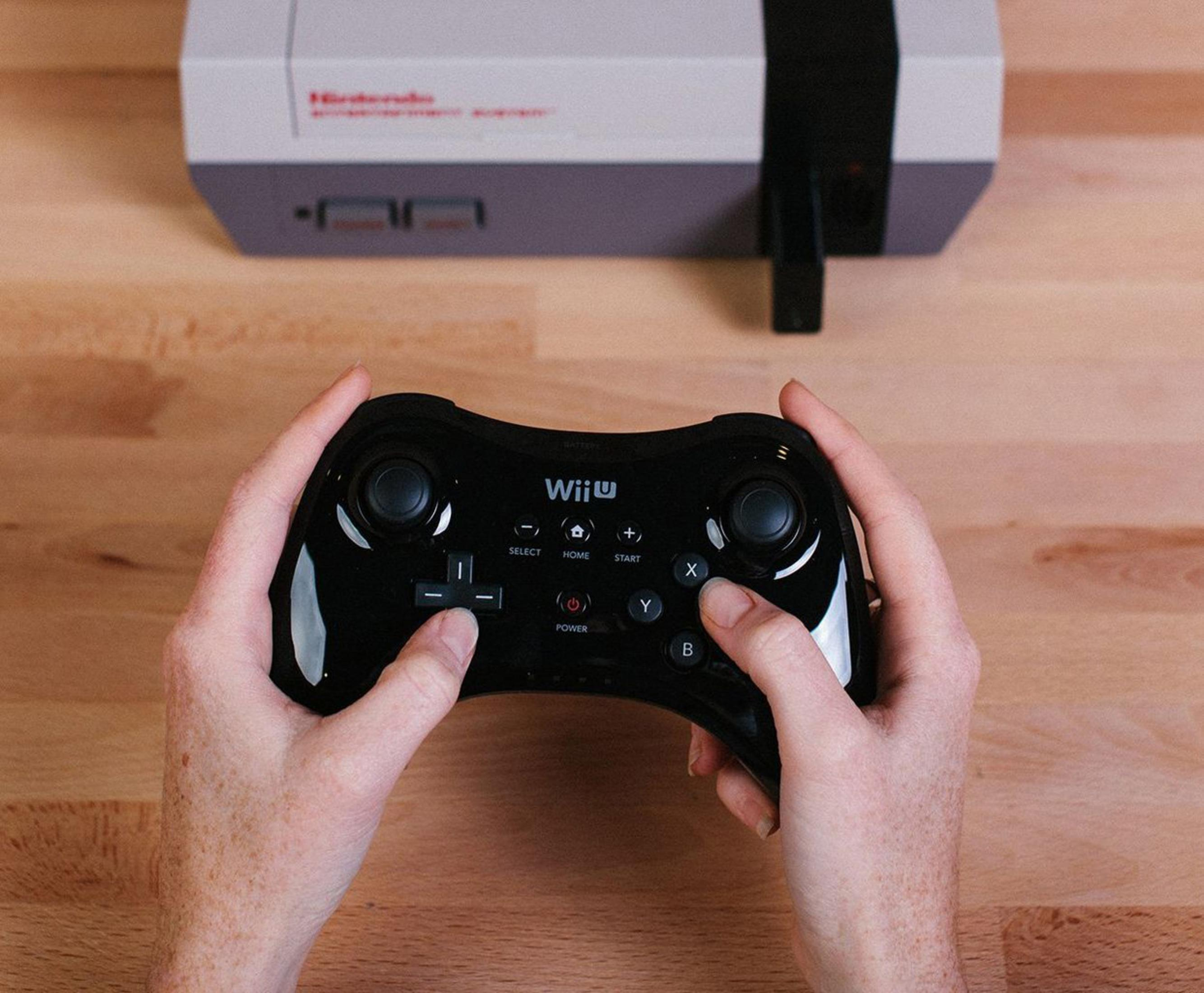 Osoba trzyma czarny kontroler Wii U, w tle konsola NES.