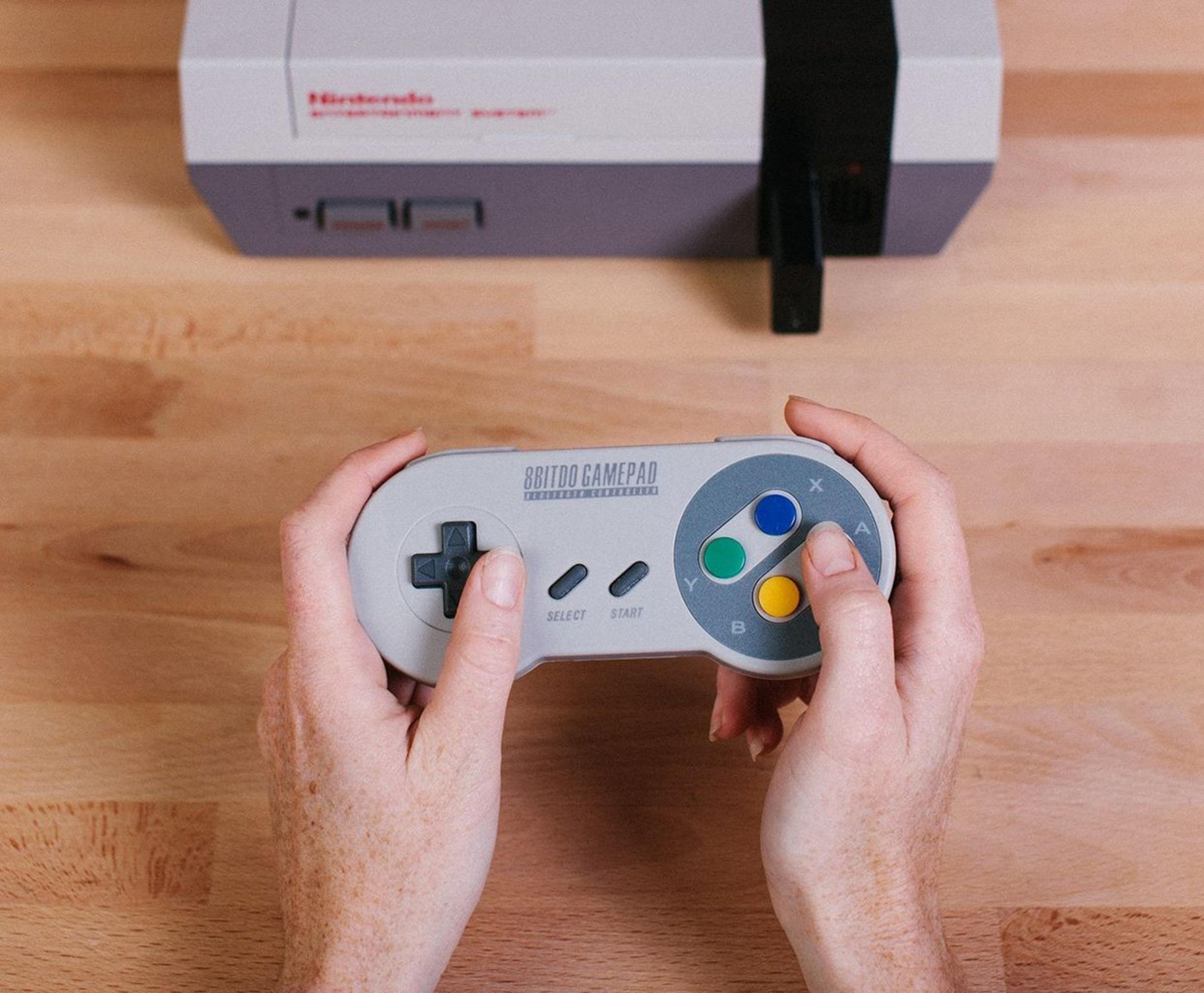 Dłonie trzymają szary kontroler przed konsolą Nintendo NES.