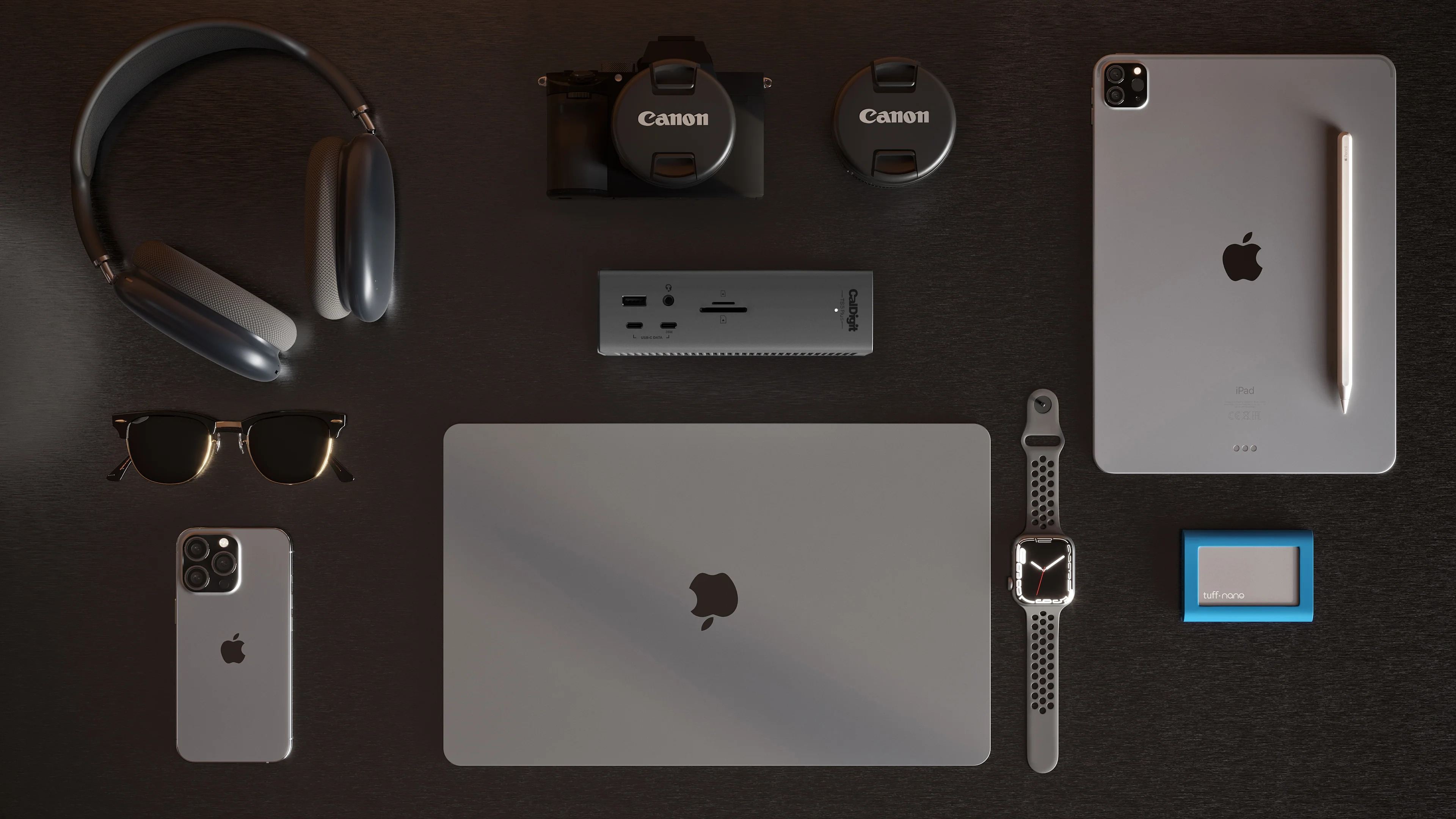 Szare słuchawki, okulary przeciwsłoneczne, iPhone, laptop, aparat, stacja dokująca, iPad, długopis, zegarek, niebieskie etui.