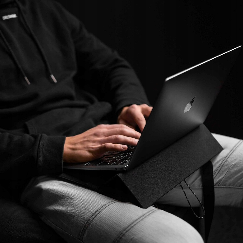 Osoba pisze na czarnym laptopie na kolanach, ciemne tło.