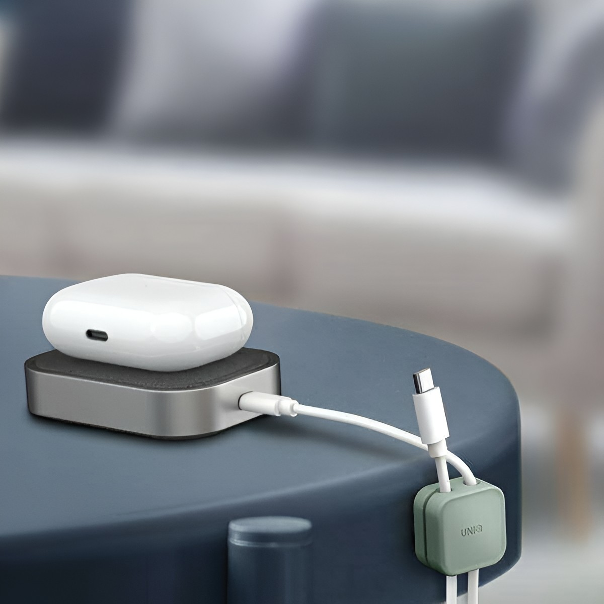 Białe etui słuchawek na ładowarce, podłączone białym kablem USB-C, na niebieskim stole.