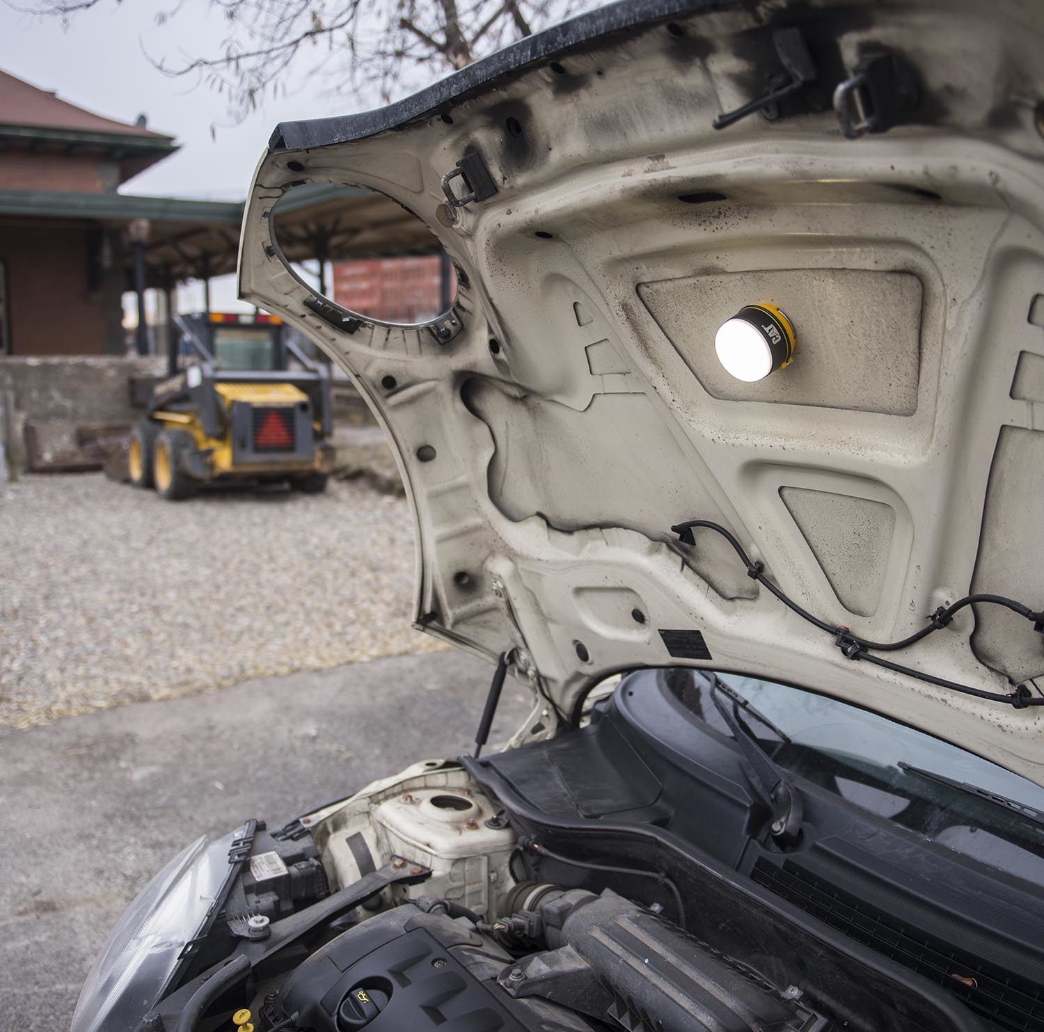 Una luz brilla bajo el capó abierto de un coche. Un minicargador está al fondo.