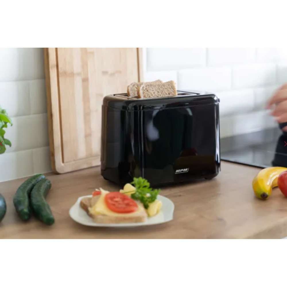 Tostapane nero con pane, accanto un sandwich, cetrioli e frutta su un tavolo di legno.