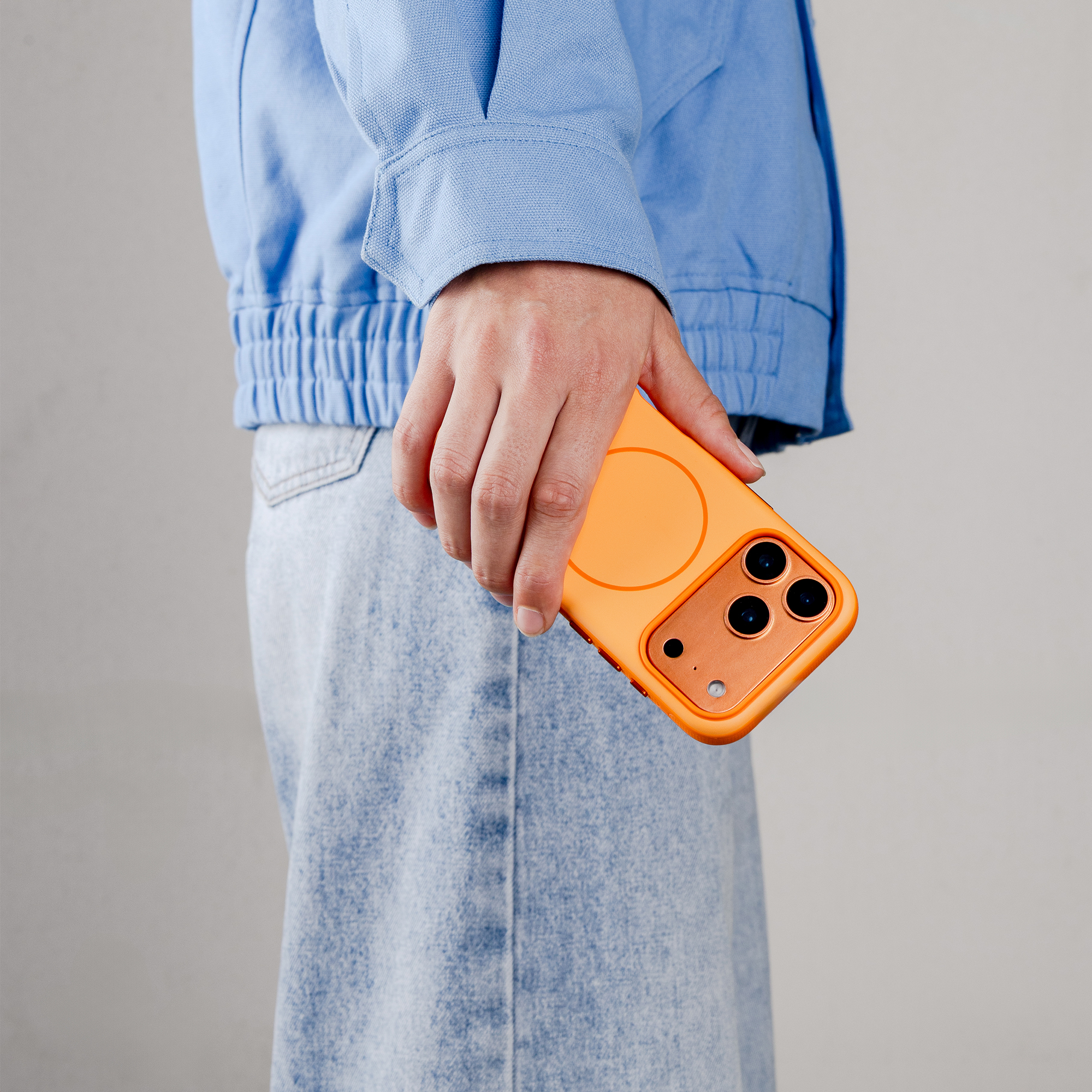 Een hand houdt een oranje telefoonhoesje met drie camera's vast. Persoon draagt blauwe jas en spijkerbroek.