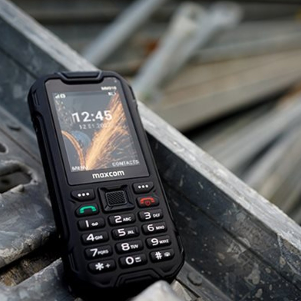 Czarny telefon komórkowy Maxcom leży na metalowych belkach. Wyświetlany czas to 12:45.