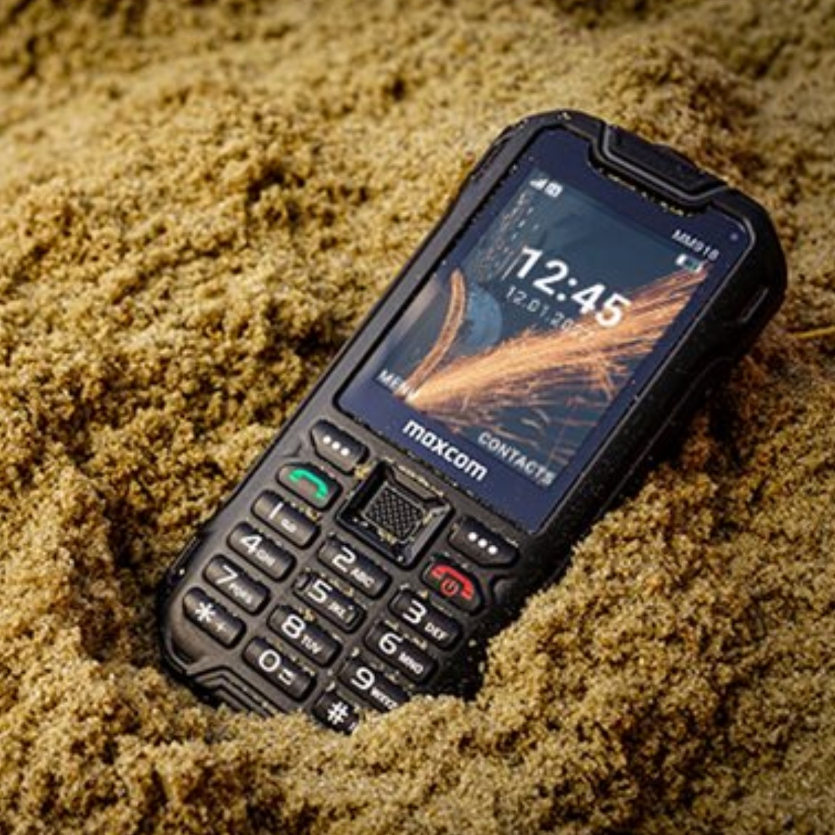 Telefon Maxcom leży w piasku. Godzina to 12:45.