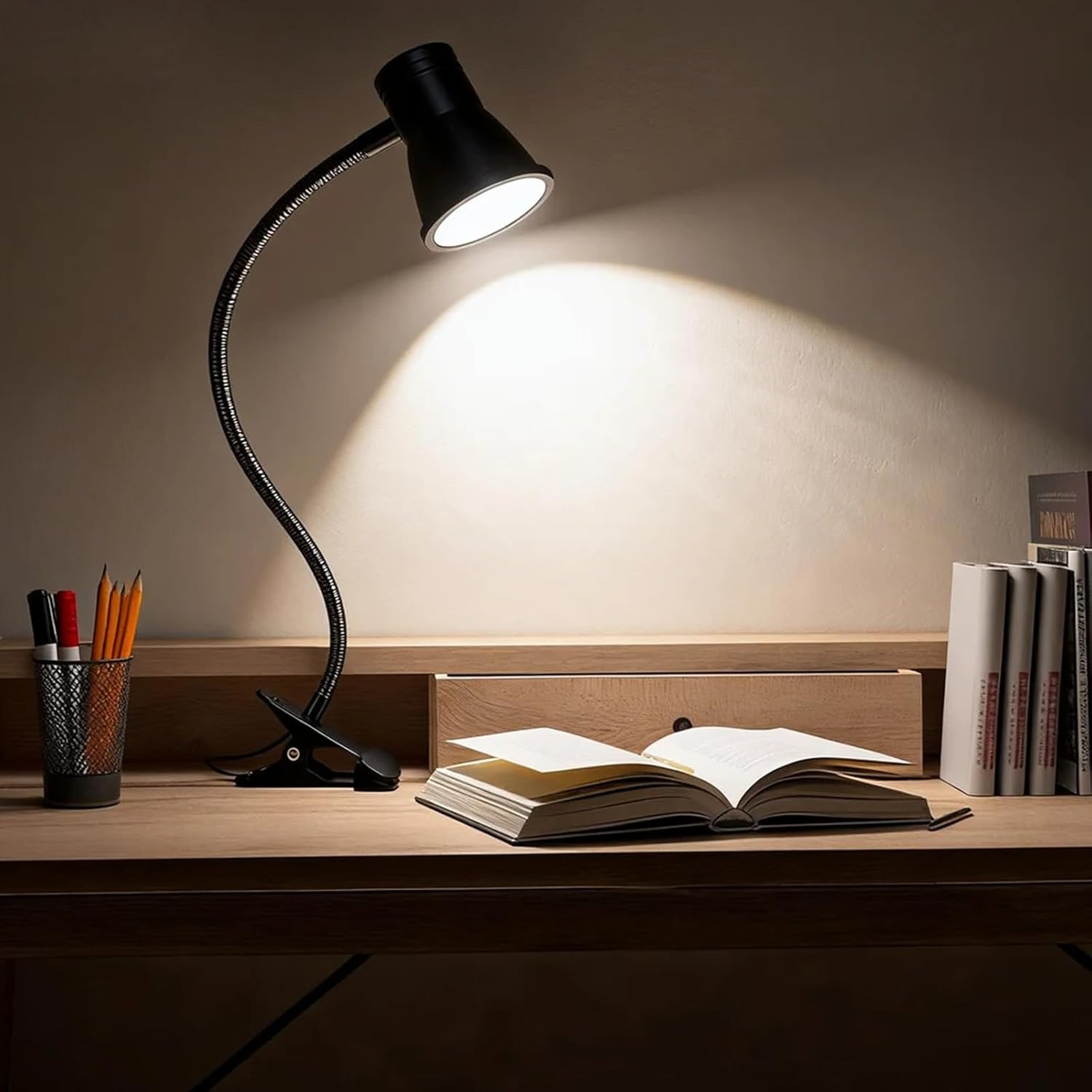 Eine schwarze Klemmleuchte beleuchtet ein offenes Buch auf einem Holzschreibtisch mit Stiften und Büchern.