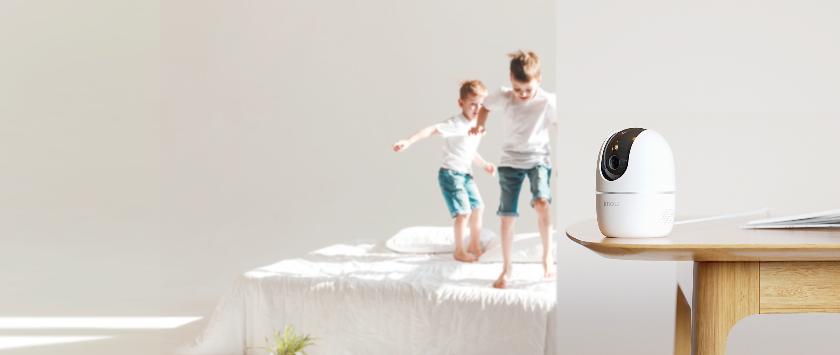 Una cámara de seguridad blanca sobre una mesa de madera, con dos niños borrosos saltando en una cama al fondo.