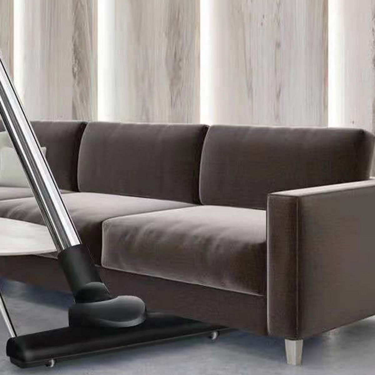Ein Staubsaugerkopf reinigt vor einem braunen Sofa mit hellen Vorhängen.