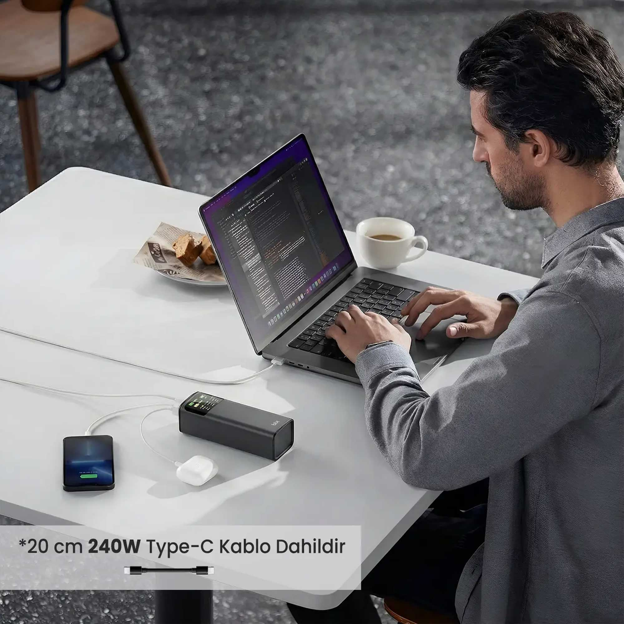 Adam dizüstü bilgisayarda yazıyor, şarj kablosu telefon, powerbank, kulaklık bağlı.
