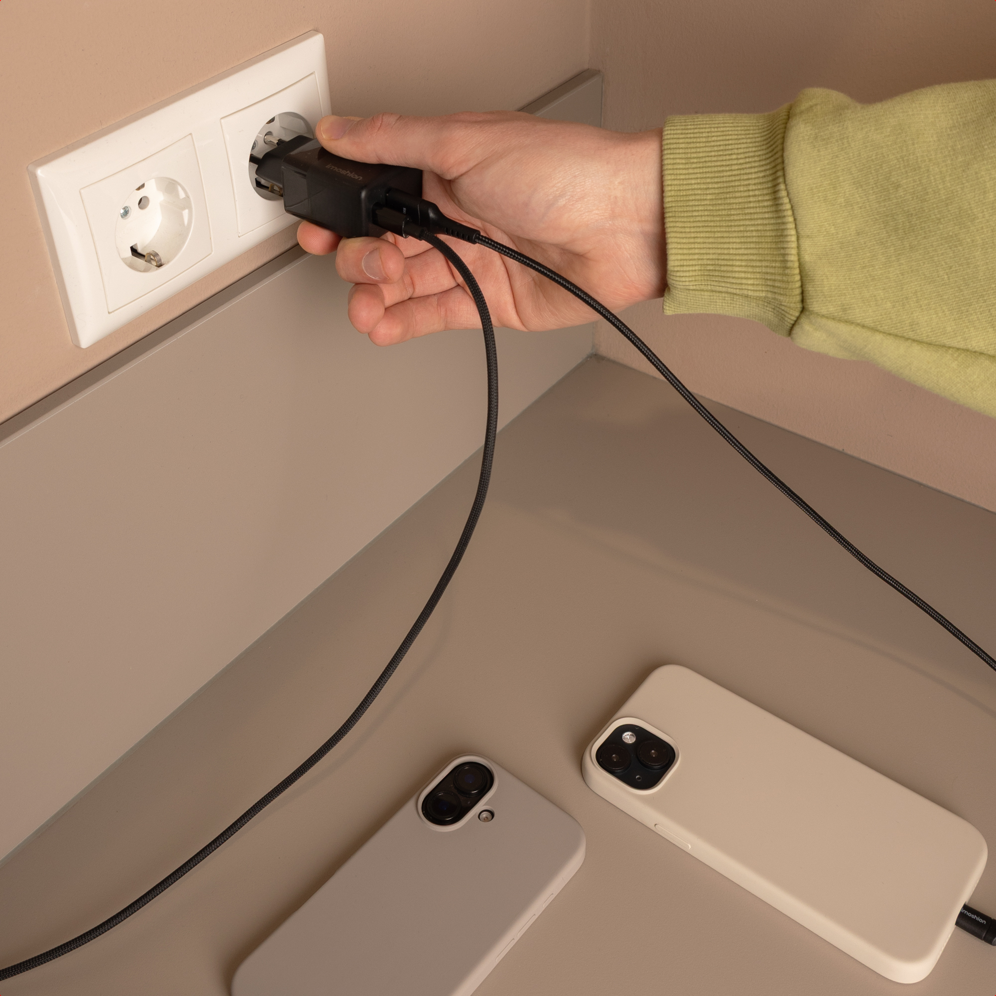 Eine Hand steckt einen schwarzen Netzstecker in eine weiße Steckdose. Zwei Handys liegen daneben.