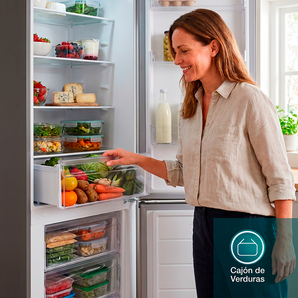 Una mujer abre un cajón de verduras en un refrigerador abierto lleno de comida.