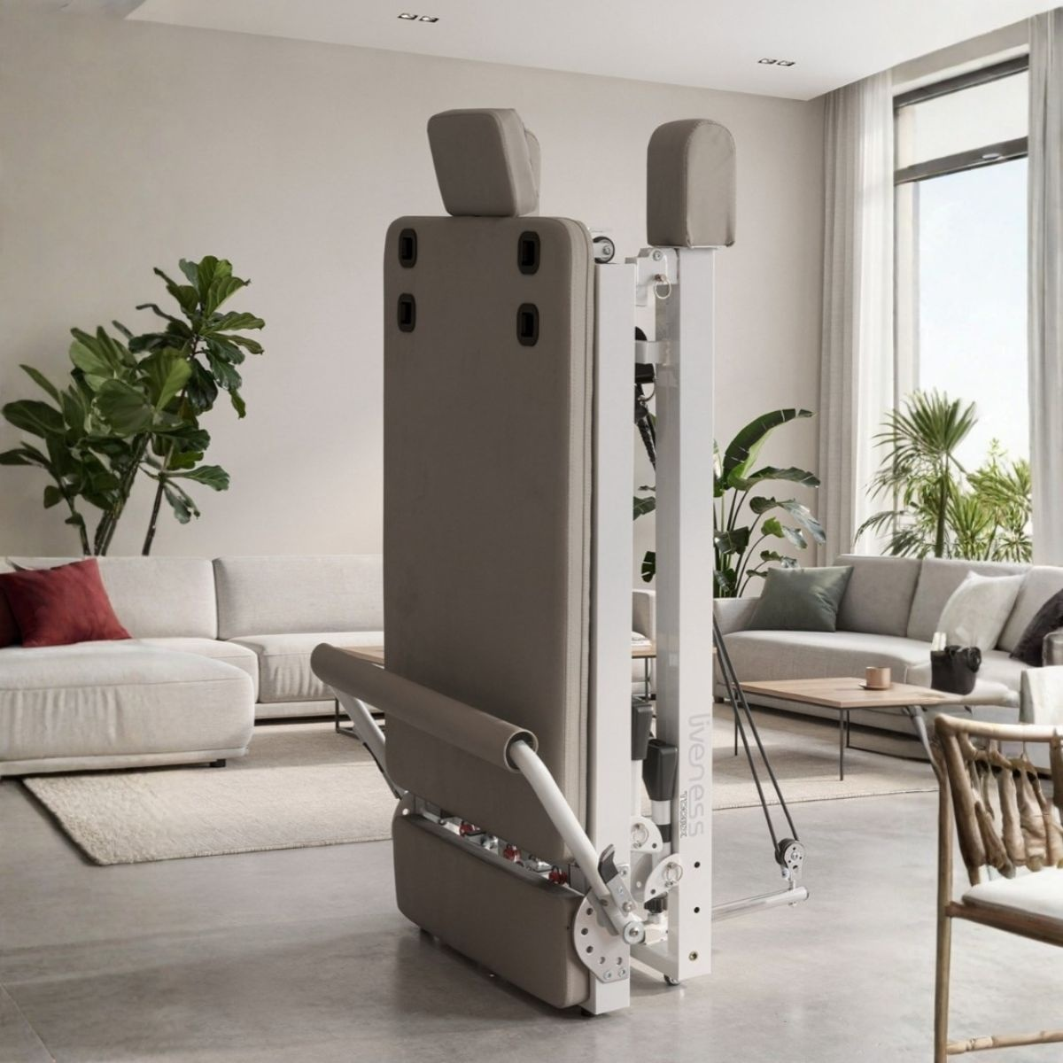 A folded grey Pilates machine stands in a bright living room with sofas and plants.