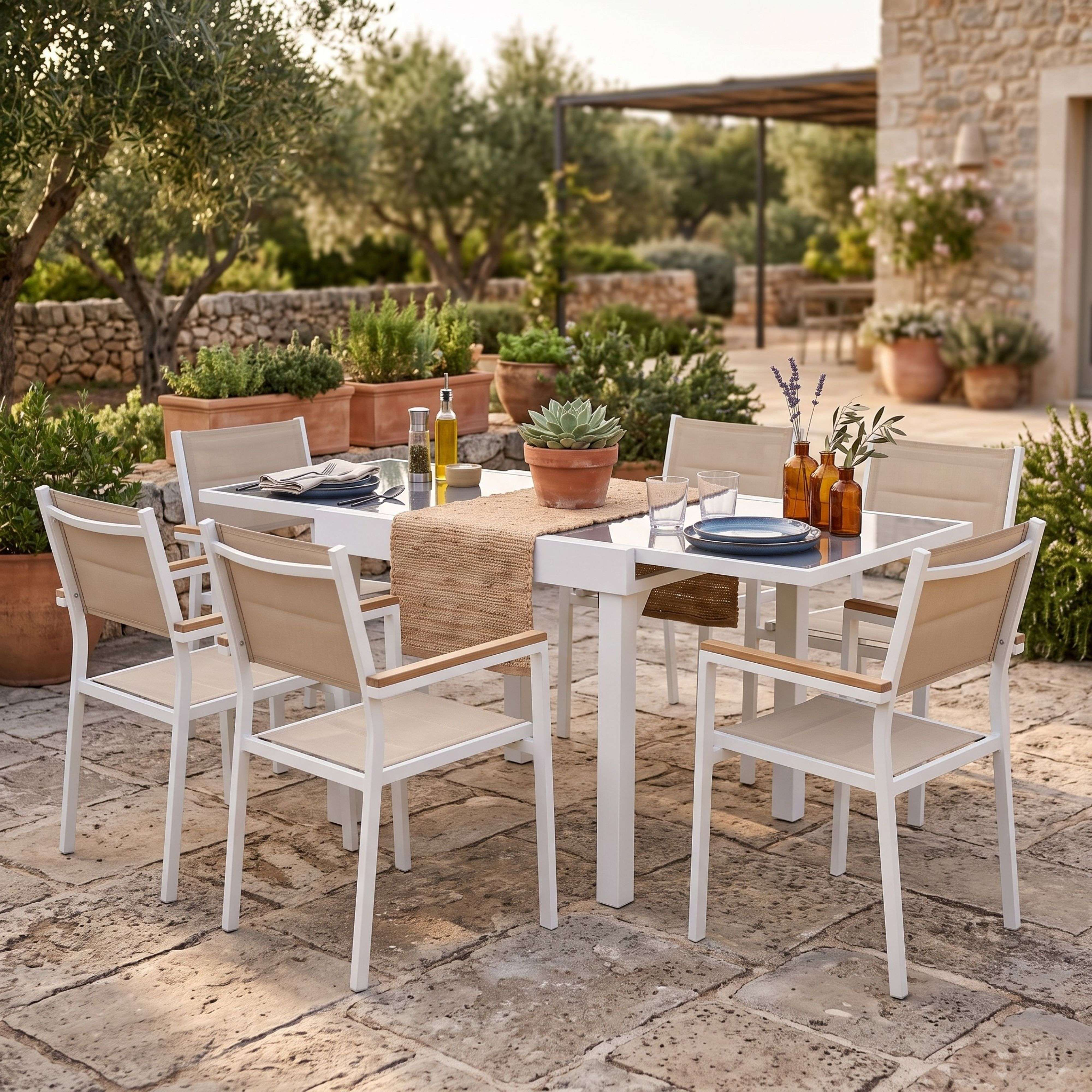 Mesa exterior puesta con sillas blancas, rodeada de plantas y muros de piedra.