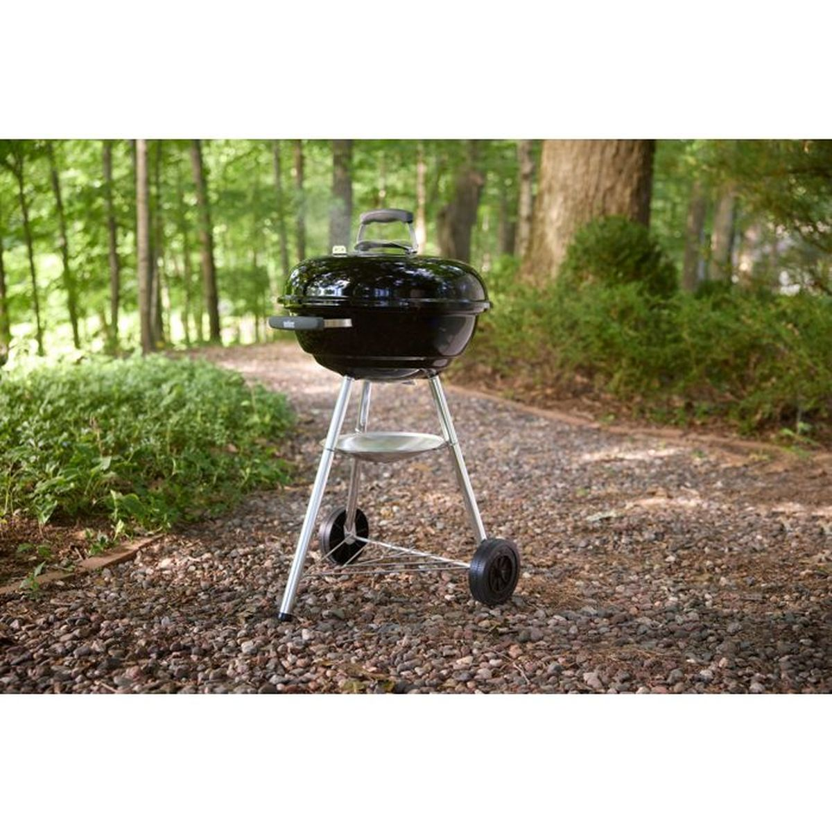 Ein schwarzer Holzkohlegrill mit Deckel steht auf einem Kiesweg im Wald.