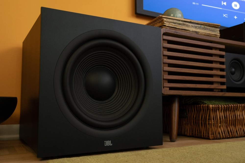 Czarny głośnik subwoofer JBL na żółtym dywanie przed drewnianą szafką RTV.